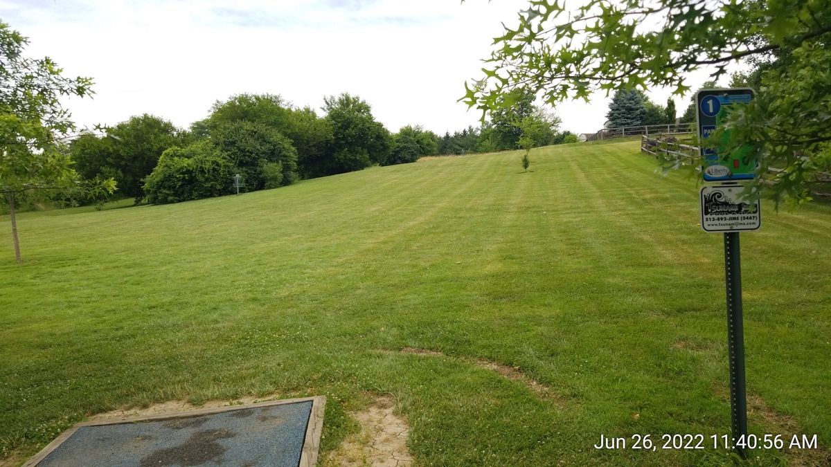 Tee pad of Hole 1, with basket in the left hand corner by bushes.  Camera angle is pointed towards Hole 2 basket/Hole 3 tee pad in top right corner