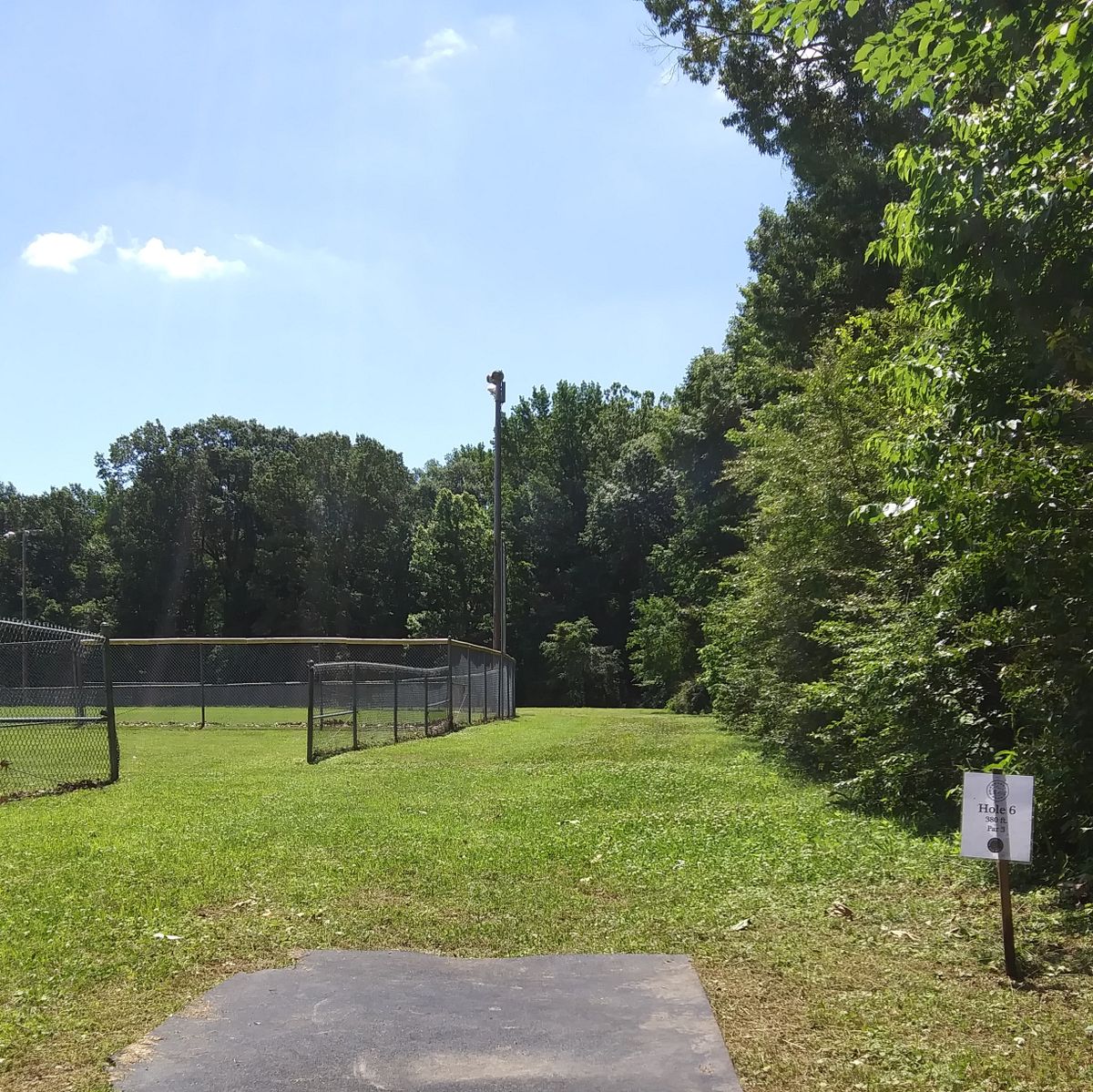 Hole 6's Tee pad. The basket is past the light pole on the left in an opening outside of the ball field's fence.