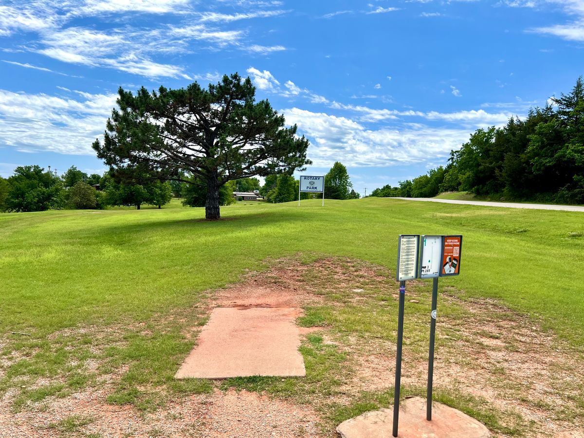 Hole 1. 285 ft par 3 with a tree and sign in the path and a public road along the right