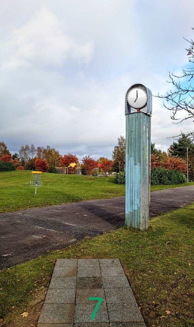 Hål 7 Gröna. Its discgolf  o'clock Hole7, 77m
 Mando-skyllt (grön) till vänster. DZ markerad med en marksten placerad till höger om skyllten. OB till höger och framför korg. Asfalt och stenlagda ytor utgör OB på banan (tees borträknat) Stenringen runt korgen är ej i spel på detta hål.